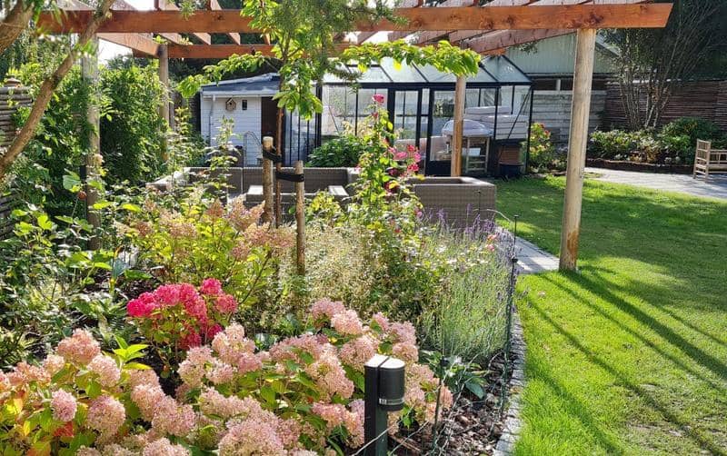 Komplet haveanlæg med træpergola, blomstrende staudebed og tæt græsplæne ved drivhus – eksempel fra Sjælland